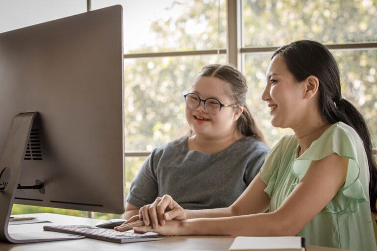 Portrait shot of happy young down syndrome daughter and mother smiling look at camera while learning online via computer.