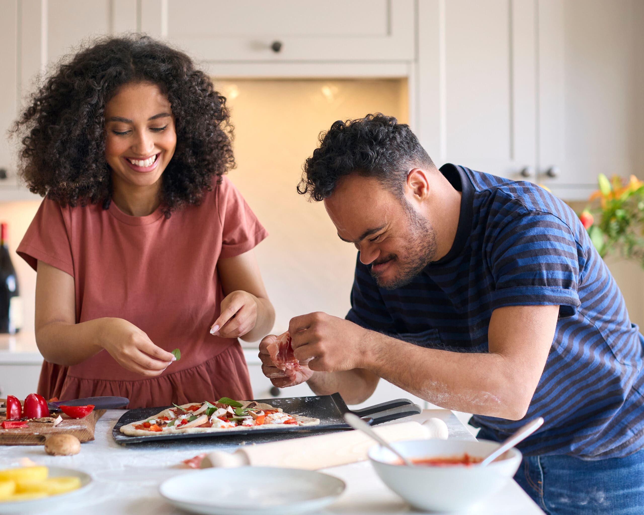 Couple At Home With Man With Down Syndrome And Woman Putting Toppings On Pizza In Kitchen Together