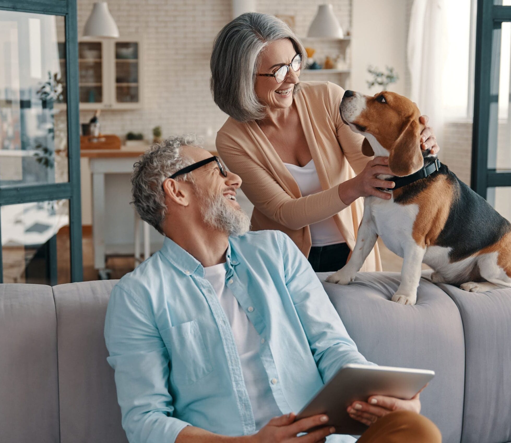 An older couple pet their beagle on the sofa while looking at an electronic tablet