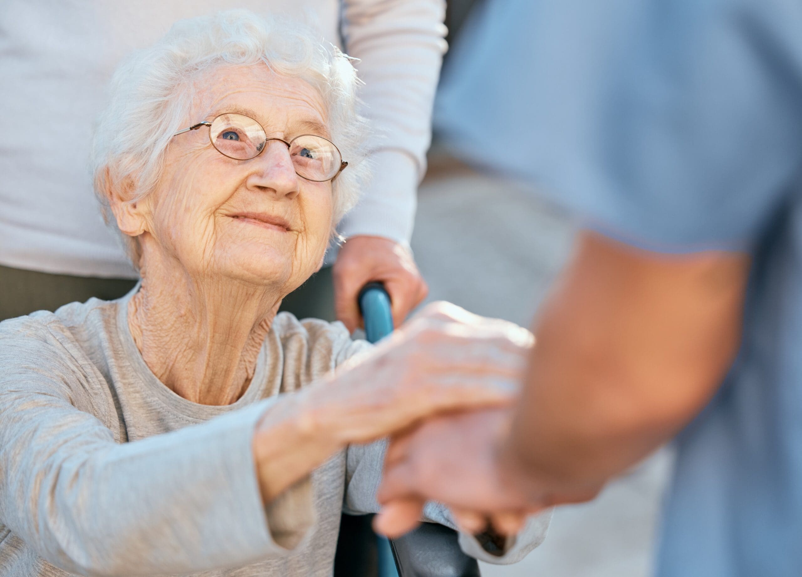 An elderly woman holds her carers hand while someone pushes her wheelchair