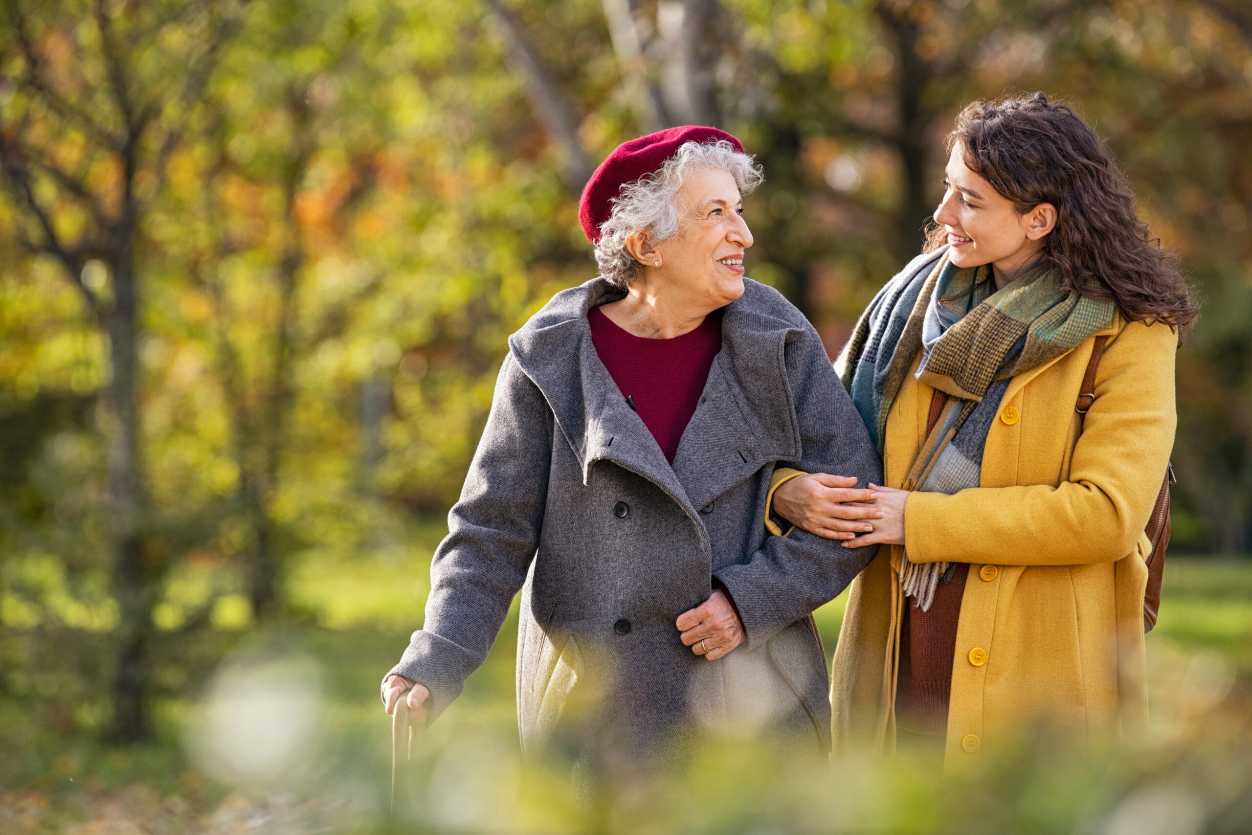 An elderly woman in a red hat and a younger woman in a yellow coat walk arm in arm